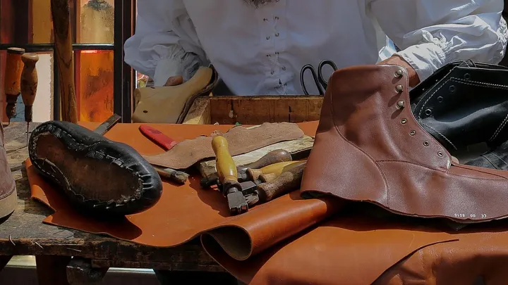 Mesa de zapatero con una bota de cuero marrón a medio montar, una suela cosida sobre su horma, pinzas de montar, lezna y cuero en rollo