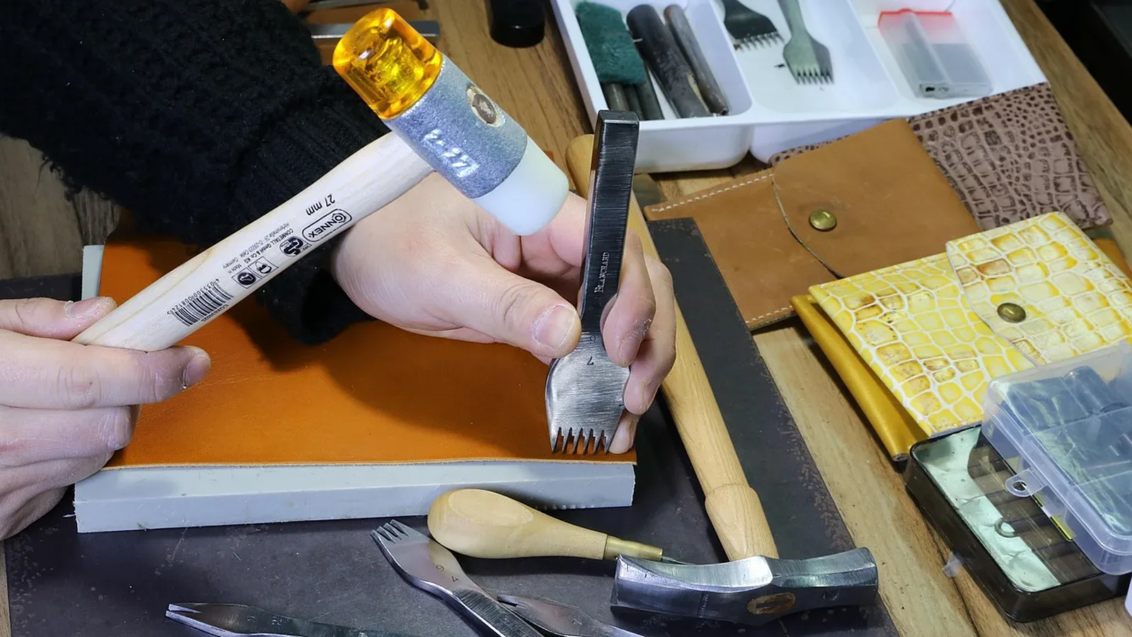 Manos marcando con mazo y pinza de coser sobre un cuero color suela, con recortes de cuero liso y estampado alrededor en la mesa de taller